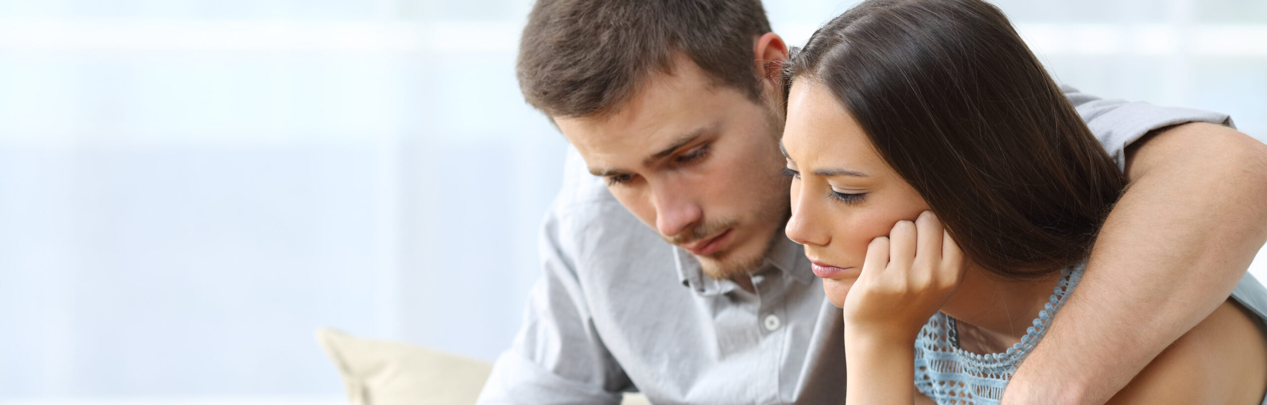 Sad couple comforting each other sitting on a couch in the living room at home due to recurrent miscarriage | Arizona Reproductive Medicine Specialists